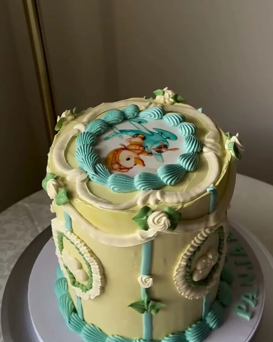 A pale yellow buttercream cake with mint piping and a hand painted illustrated plaque on top